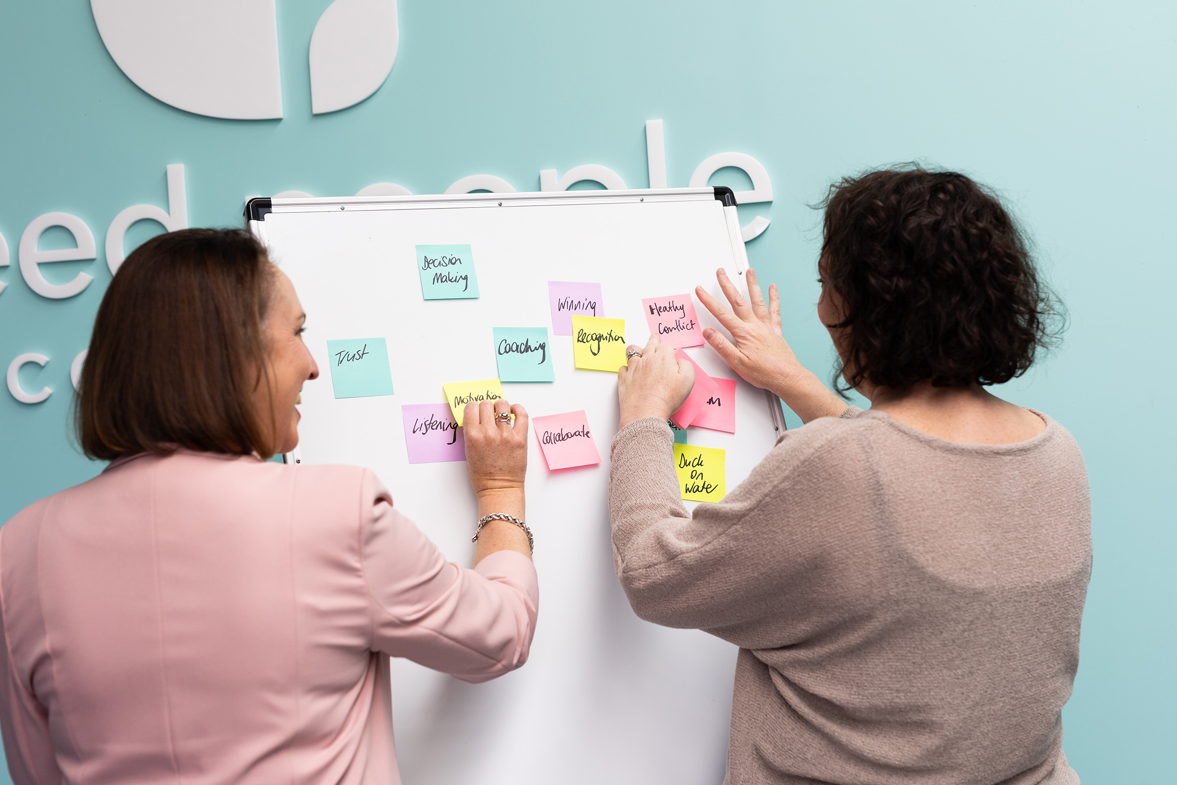 Two Seed consultants writing on sticky notes during a culture mapping workshop in front of the Seed People branded wall.