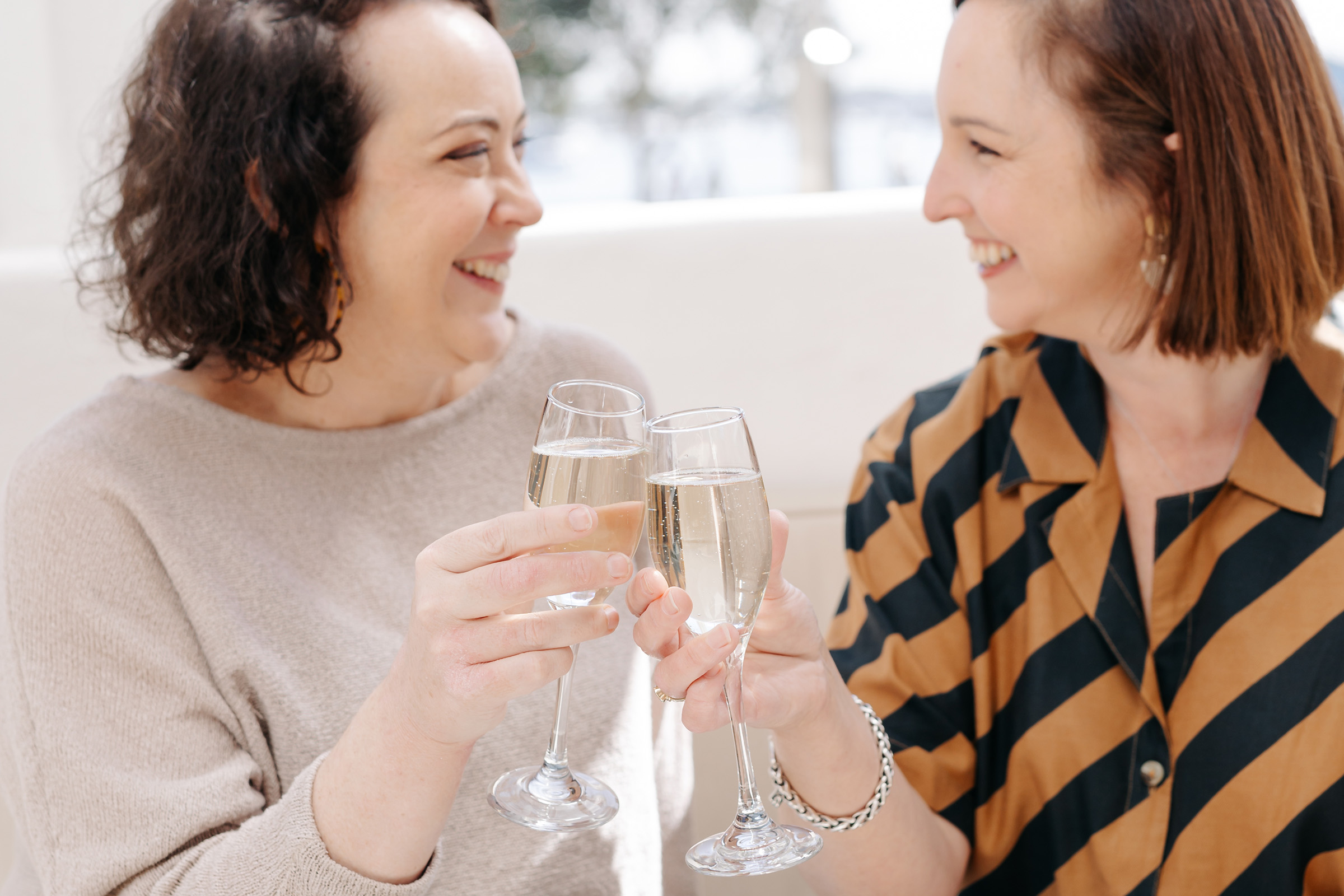 Stacey Kelly and Julia Fiore clinking champagne glasses while laughing together in a bright room.