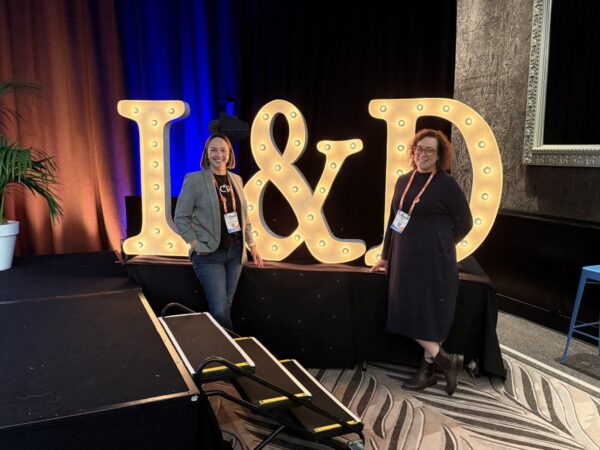 Stacey Kelly and Julia Fiore of Seed People Consulting at the 2026 HR and L&D Innovation and Tech Fest in Auckland, standing beside a large light-up 'L&D' marquee sign.